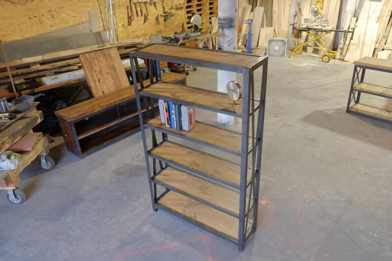 May include: A black metal bookshelf with five wooden shelves. The shelves are made of reclaimed wood and have a rustic look. The bookshelf is standing on a concrete floor in a workshop.