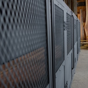 May include: A row of dark gray metal cabinets with diamond mesh and solid panels. The cabinets have a utilitarian design with visible hinges and handles. The background shows wood planks and a concrete floor, suggesting a workshop or storage area.