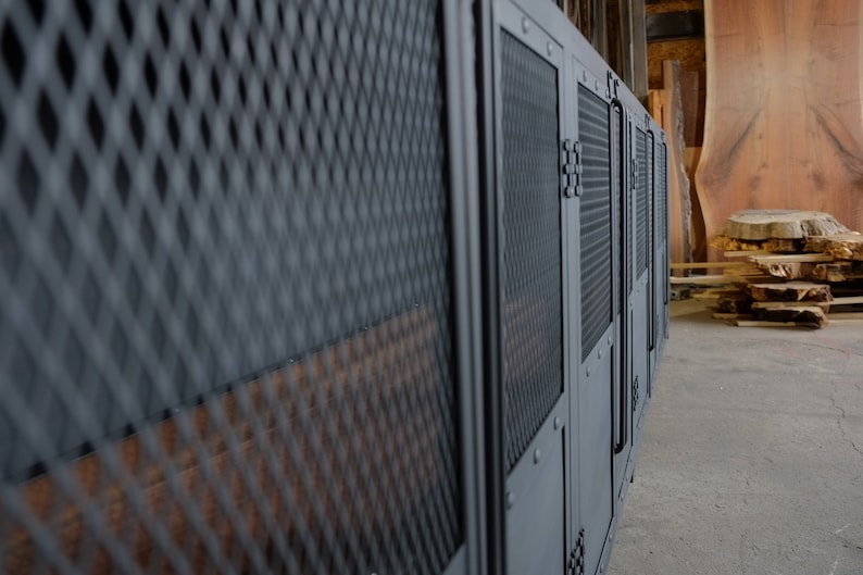 May include: A row of dark gray metal cabinets with diamond mesh and solid panels. The cabinets have a utilitarian design with visible hinges and handles. The background shows wood planks and a concrete floor, suggesting a workshop or storage area.
