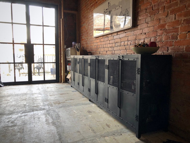 May include: A long, black metal cabinet with multiple doors and mesh inserts. The cabinet is in an industrial-style room with a brick wall and a large window. A bowl of decorative items sits on top.