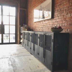 May include: A long, black metal cabinet with multiple doors and mesh panels. The cabinet is set against a red brick wall and sits on a concrete floor. A framed picture hangs above the cabinet, and a bowl of fruit rests on top.