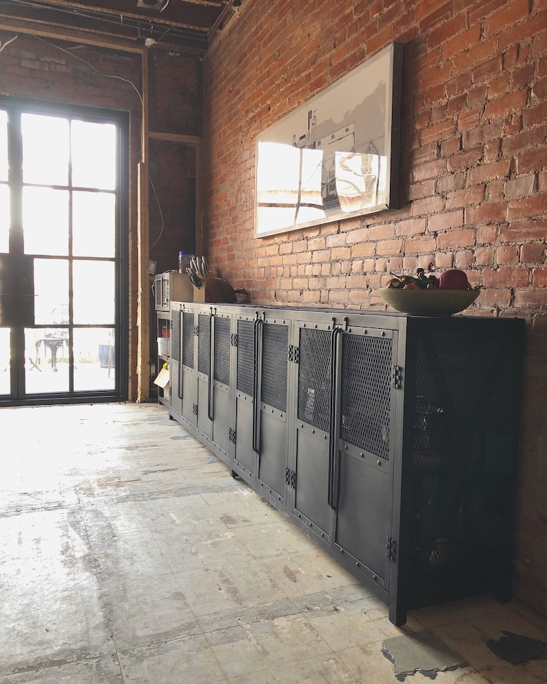 May include: A long, black metal cabinet with multiple doors and mesh panels. The cabinet is set against a red brick wall and sits on a concrete floor. A framed picture hangs above the cabinet, and a bowl of fruit rests on top.