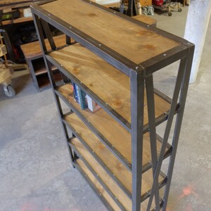May include: A five-shelf industrial style bookcase with a black metal frame and wooden shelves. The shelves are made of natural wood with a light brown stain. The bookcase has a simple design with a slight angle on the front legs.