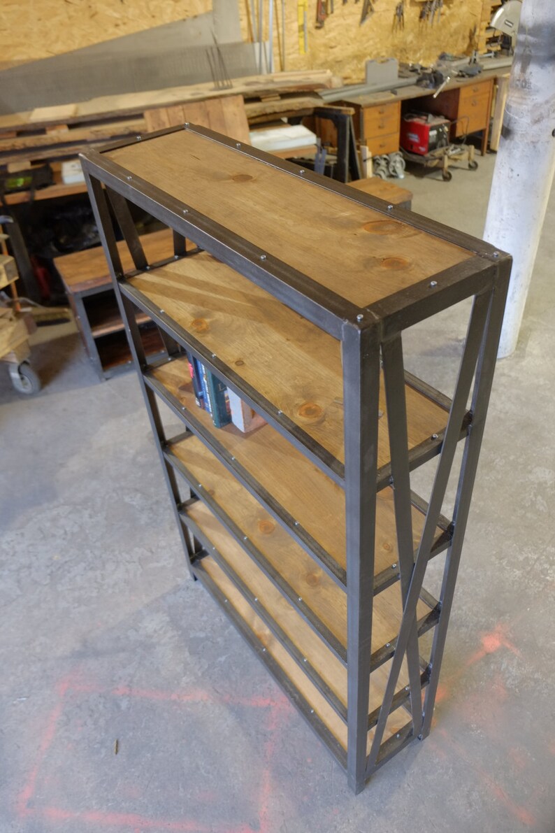 May include: A five-shelf industrial style bookcase with a black metal frame and wooden shelves. The shelves are made of natural wood with a light brown stain. The bookcase has a simple design with a slight angle on the front legs.