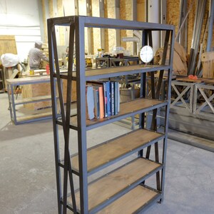 May include: A black metal industrial style bookshelf with five shelves. The shelves are made of wood and have a rustic finish. The bookshelf is standing in a workshop setting.