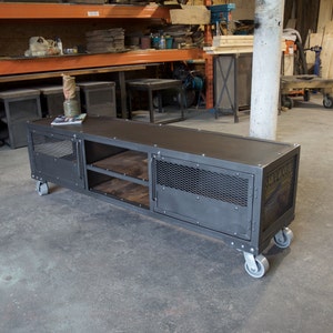 May include: A black metal industrial style media console with two doors, two shelves, and four wheels. The console has a distressed finish and a mesh design on the doors. The console is sitting on a concrete floor.