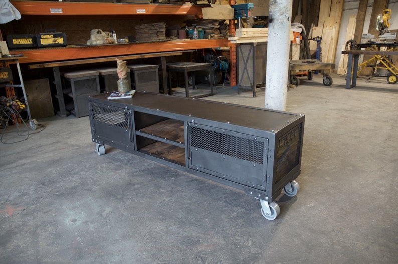 May include: A black metal industrial style media console with two doors, two shelves, and four wheels. The console has a distressed finish and a mesh design on the doors. The console is sitting on a concrete floor.