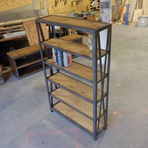May include: A black metal bookshelf with five wooden shelves. The shelves are made of reclaimed wood and have a rustic look. The bookshelf is standing on a concrete floor.