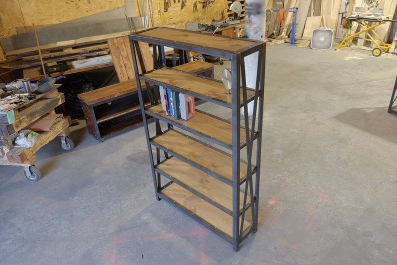 May include: A black metal bookshelf with five wooden shelves. The shelves are made of reclaimed wood and have a rustic look. The bookshelf is standing on a concrete floor.