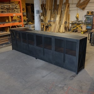 May include: A large, black metal cabinet with a rectangular shape. The cabinet has multiple door panels with a diamond-patterned mesh design. The cabinet is in a workshop setting, with wood and tools visible in the background.