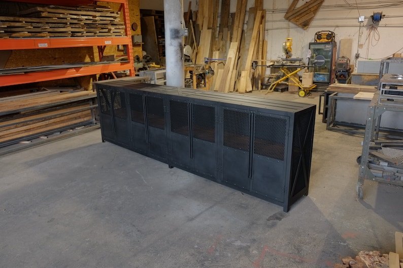 May include: A large, black metal cabinet with a rectangular shape. The cabinet has multiple door panels with a diamond-patterned mesh design. The cabinet is in a workshop setting, with wood and tools visible in the background.