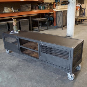 May include: A gray metal industrial style TV stand with two doors, two shelves, and four wheels. The stand has a mesh design on the doors and a wood top.