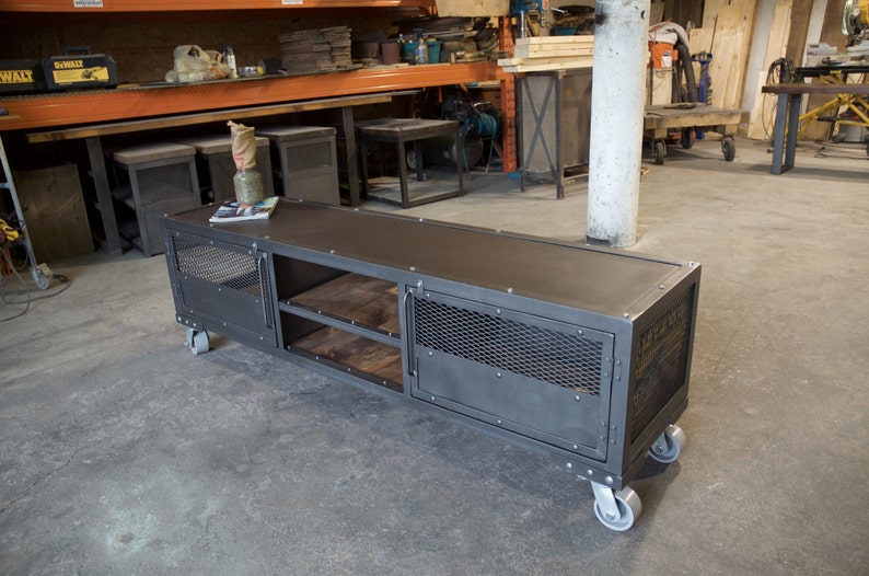 May include: A gray metal industrial style TV stand with two doors, two shelves, and four wheels. The stand has a mesh design on the doors and a wood top.