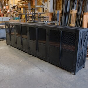 May include: A long, black metal cabinet with mesh door panels and X-shaped supports on the ends. The cabinet has multiple doors and a flat top, suitable for storage or display. The industrial design is visible.