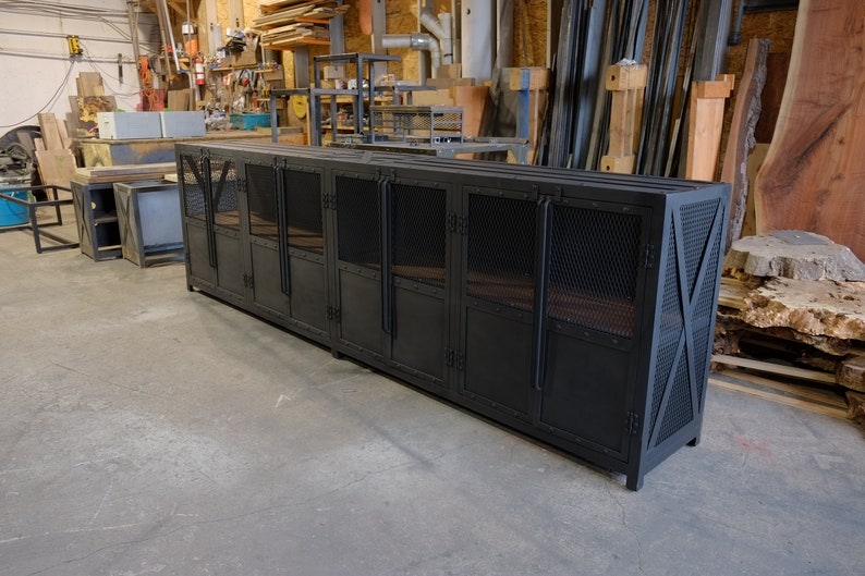 May include: A long, black metal cabinet with mesh door panels and X-shaped supports on the ends. The cabinet has multiple doors and a flat top, suitable for storage or display. The industrial design is visible.