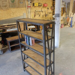 May include: A black metal bookshelf with five wooden shelves. The shelves are made of reclaimed wood and have a rustic look. The bookshelf is standing on a concrete floor.