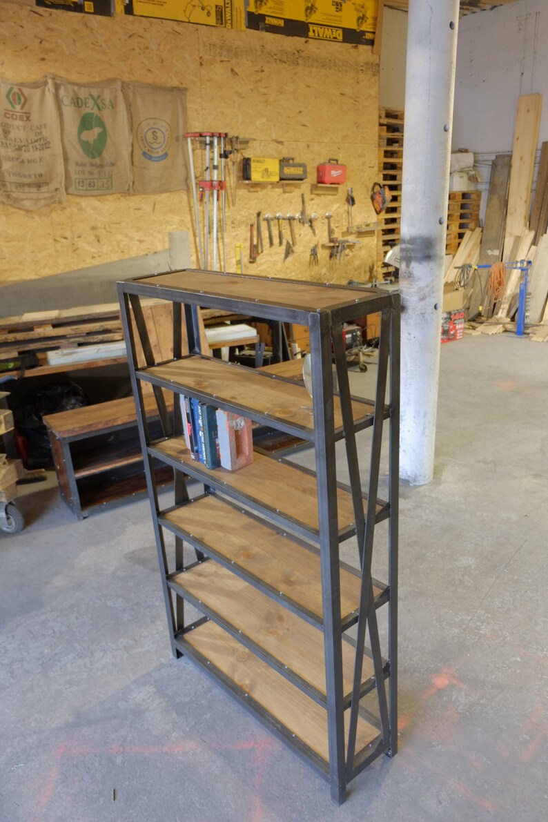 May include: A black metal bookshelf with five wooden shelves. The shelves are made of reclaimed wood and have a rustic look. The bookshelf is standing on a concrete floor.