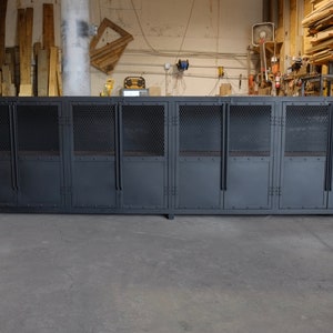 May include: A large, industrial-style cabinet with multiple compartments. The cabinet is painted a dark gray and features metal mesh doors and solid metal panels. The doors have handles and are mounted on hinges. The cabinet is set on a low, rectangular base.