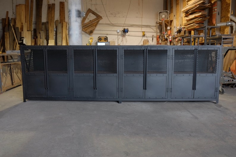May include: A large, industrial-style cabinet with multiple compartments. The cabinet is painted a dark gray and features metal mesh doors and solid metal panels. The doors have handles and are mounted on hinges. The cabinet is set on a low, rectangular base.