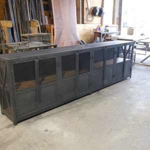 May include: A long, dark gray metal cabinet with multiple doors and mesh panels. The cabinet has a rectangular shape and is likely used for storage. The doors have a latch closure. The cabinet is in a workshop setting.