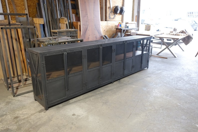 May include: A long, dark gray metal cabinet with multiple doors and mesh panels. The cabinet has a rectangular shape and is likely used for storage. The doors have a latch closure. The cabinet is in a workshop setting.