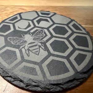 Puede incluir: Un posavasos redondo de pizarra gris oscuro con un diseño grabado detallado. El diseño presenta una abeja con intrincados detalles de alas y cuerpo, sobre un patrón de panal. El posavasos tiene un borde natural y texturizado.