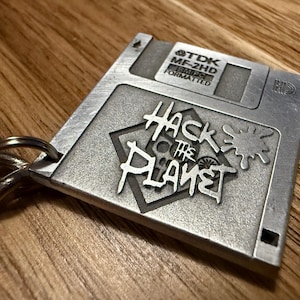 Peut inclure: Un porte-clés en métal argenté en forme de disquette avec le texte "Hack the Planet" et "TDK MF-2HD 1.44MB Formatted" dessus.