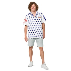 May include: A white patterned shirt with a blue design, paired with light green shorts and white trainers. The shirt features a camp collar and short sleeves. The shirt has a red logo on the left sleeve.