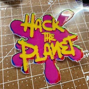 Op de afbeelding: Een kleurrijke sticker met de tekst "HACK THE PLANET" in het geel, omlijnd met rood, blauw en groen. De tekst staat op een roze, onregelmatig gevormde achtergrond.