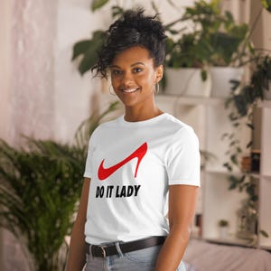 Könnte beinhalten: Weißes T-Shirt mit einem Design eines roten High-Heel-Schuhs und dem Text "DO IT LADY" in Schwarz. Das T-Shirt hat einen Rundhalsausschnitt.