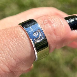 Puede incluir: Un anillo de plata con una banda texturizada y un pequeño diseño grabado de un planeta con anillos. El anillo se lleva en un dedo.