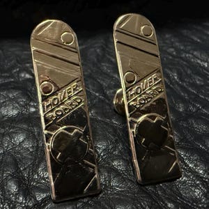 May include: Two gold-colored metal hoverboard keychains with the words "Hover Board" engraved on them. Each keychain has a circular design with a cross in the center.