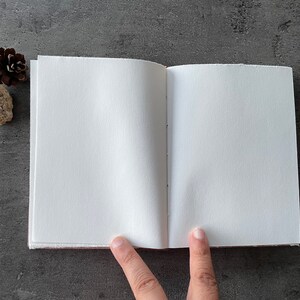 May include: A blank, white, paper notebook with a brown cover is open to a clean, white page. The notebook is lying flat on a gray surface.
