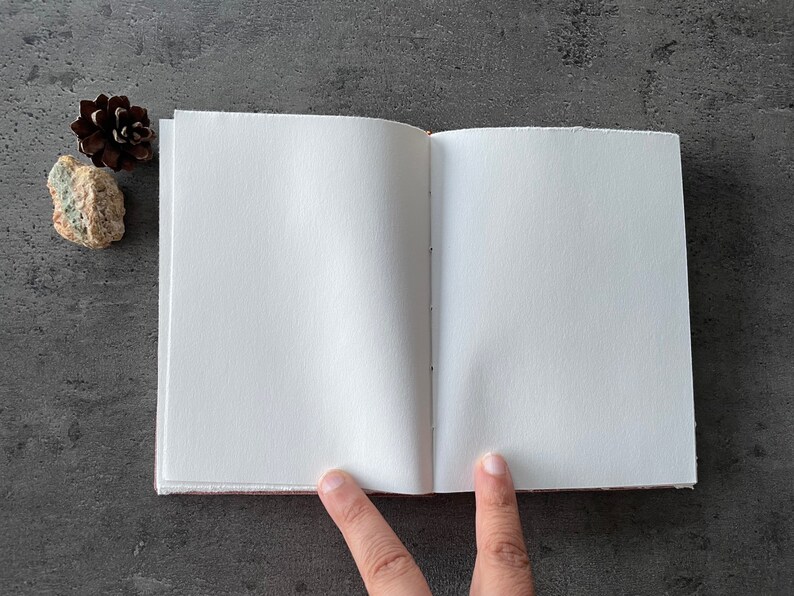 May include: A blank, white, paper notebook with a brown cover is open to a clean, white page. The notebook is lying flat on a gray surface.