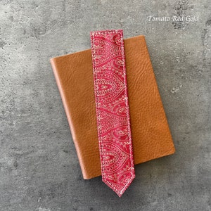 Może przedstawiać: Zakładka w kolorze czerwonym i złotym z wzorem paisley i białymi szwami. Zakładka leży na brązowym sk&oacute;rzanym notesie. Tekst "Tomato Red Gold" jest widoczny w prawym g&oacute;rnym rogu obrazu.