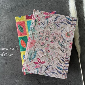 May include: Two notebooks with hard covers. The notebook on the left has a pink, yellow, and green checkered pattern. The notebook on the right has a pink and white floral pattern with a textural surface. The text "Pink Leaves - Silk Hard Cover" is visible on the image.
