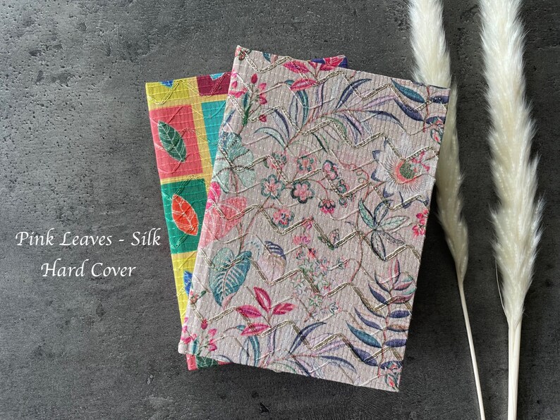 May include: Two notebooks with hard covers. The notebook on the left has a pink, yellow, and green checkered pattern. The notebook on the right has a pink and white floral pattern with a textural surface. The text "Pink Leaves - Silk Hard Cover" is visible on the image.