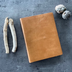 May include: A closed, tan leather journal rests on a grey surface, accompanied by two pieces of driftwood and two small, textured, grey-green stones. The journal has a smooth, rectangular cover, suggesting a classic design. The natural elements add a rustic touch.