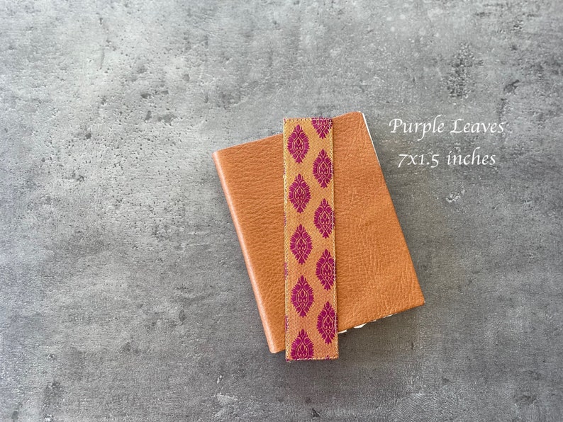 Może przedstawiać: Brązowy sk&oacute;rzany notes z zakładką w kolorze złotym i fioletowym. Notes ma wymiary 17,78 x 3,81 cm i zawiera napis "Purple Leaves" na okładce.