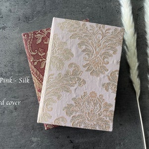 May include: Two hard cover journals with a blush pink silk cover and a gold and white damask pattern cover. The text "Blush Pink - Silk" and "Hard cover" is visible on the image.