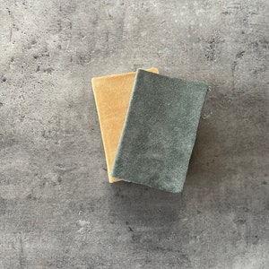 May include: Two rectangular sanding blocks, one is beige and the other is dark gray, are lying on a gray textured surface.