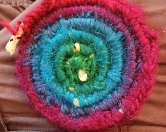 May include: A handmade, circular crochet item with a vibrant rainbow color scheme. The center is green, transitioning to blue, then purple, and finally a deep pink outer edge. The texture is clearly defined, showcasing the individual stitches.