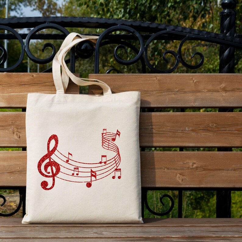 May include: A white canvas tote bag with a red glitter design of musical notes and a treble clef. The bag is sitting on a wooden bench.