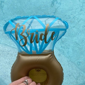 May include: A blue and gold inflatable ring shaped like a diamond ring with the word "Bride" written on it. The ring is floating in a pool of water.