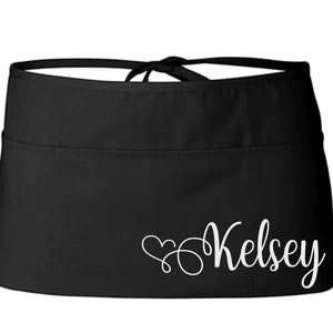 May include: Black half apron with a white heart and the name "Kelsey" written in cursive.