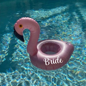May include: Pink inflatable flamingo pool float with a black beak and yellow eye, floating in a pool. The word "Bride" is written on the float. The water is a clear blue.
