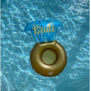 May include: A gold inflatable ring with a blue diamond-shaped top that says "Bride" in gold glitter letters. The ring is floating in a blue swimming pool.