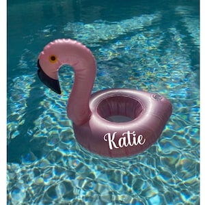 May include: Pink inflatable flamingo pool float with a cup holder and the name "Katie" printed on the side.