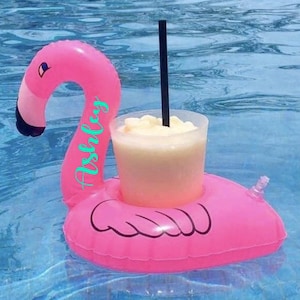 May include: Pink inflatable flamingo pool float with a clear plastic cup holder and a black straw. The flamingo has the word "Ashley" written on it in turquoise.