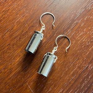 May include: A pair of silver-colored earrings. Each earring features a cylindrical design with a silver hook. The earrings are displayed on a brown wooden surface.